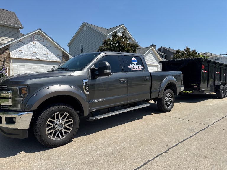 roof bros restoration branded truck 004 - Roof Bros Restoration - roofing gallery wisconsin illinois Dark gray Ford pickup truck with a Roof Bros Restoration logo decal on the door, towing a black dump trailer, parked on a...