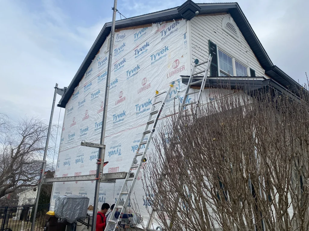 Two-story side wall wrapped in Tyvek HomeWrap with ladders and a worker preparing for siding install