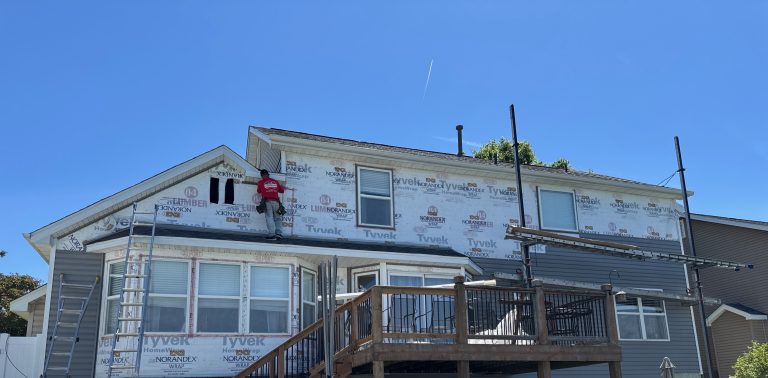 roof bros restoration siding install 001 - Roof Bros Restoration - roofing gallery wisconsin illinois A worker in a red shirt stands on a deck roof of a two-story home wrapped in Tyvek and Norandex house wrap with a ladder a...