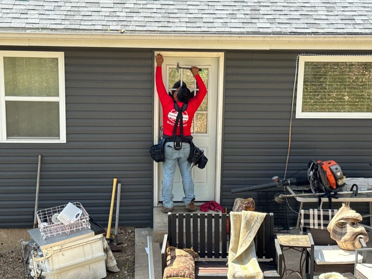roof bros restoration siding install 006 2 - Roof Bros Restoration - roofing gallery wisconsin illinois A worker in a red Roof Bros Restoration t-shirt and tool belt stands on a back patio installing or repairing trim above a...