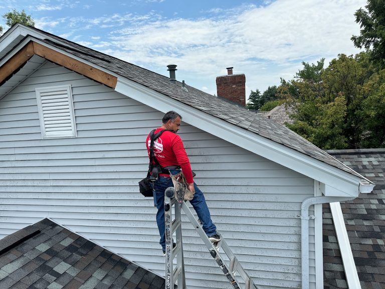 roof bros restoration siding install 007 - Roof Bros Restoration - roofing gallery wisconsin illinois A worker in a red shirt stands on a ladder against a gray-sided home holding tools near the soffit and roofline