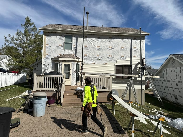 roof bros restoration siding install 015 - Roof Bros Restoration - roofing gallery wisconsin illinois A worker in a yellow-green high-visibility shirt walking across a paver patio toward a two-story residential home that has...