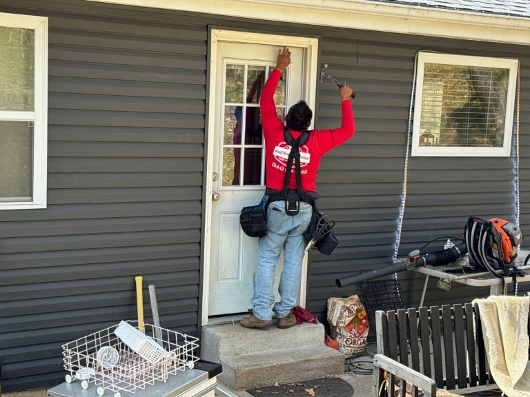 roof bros restoration siding install 017 2 - Roof Bros Restoration - roofing gallery wisconsin illinois A worker in a red Roof Bros Restoration t-shirt and tool belt stands on a concrete stoop reaching up to work on trim above...