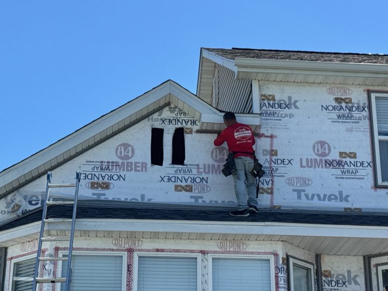 roof bros restoration siding install 018 - Roof Bros Restoration - roofing gallery wisconsin illinois A worker in a red Roof Bros Restoration shirt stands on a porch roof installing house wrap on the gable of a residential h...
