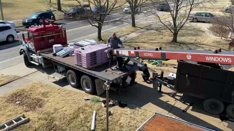 roof bros restoration video poster jobsite 015 - Roof Bros Restoration - roofing gallery wisconsin illinois Overhead angle of a red flatbed truck carrying shingle bundles next to a SIDING/WINDOWS conveyor and a dump trailer, with...